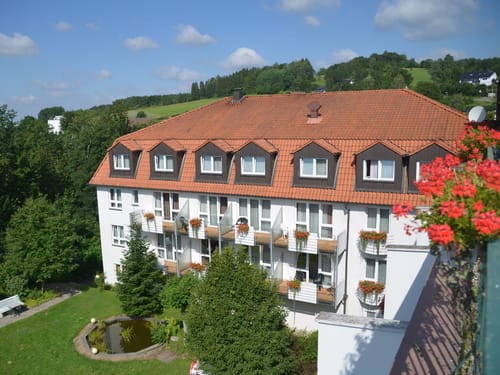 Panorama eines Hotels mit rotem Ziegeldach und blühenden Balkonpflanzen in einer grünen Landschaft.