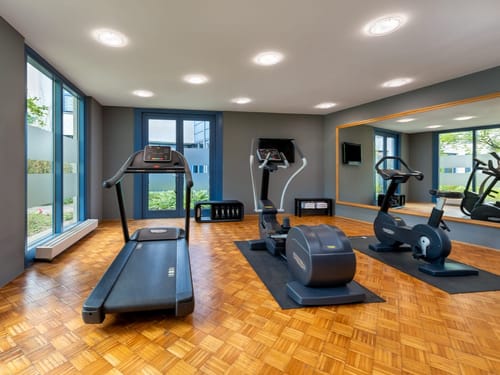 Einladender Fitnessbereich mit modernen Cardiogeräten, Parkettboden und großzügiger Verglasung für ein angenehmes Trainingserlebnis