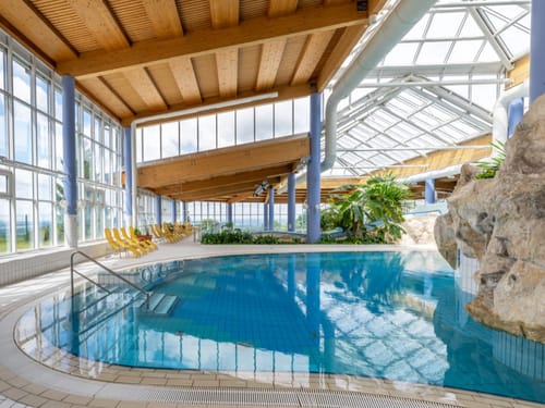 Innenraum eines modernen Hallenbads mit gläsernem Dach und blauen Liegestühlen.