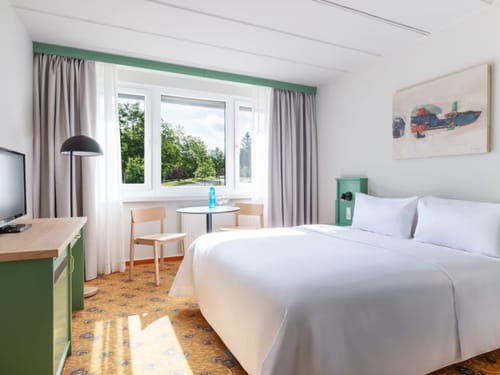 Helles und modernes Hotelzimmer mit gemütlichem Doppelbett, großem Fenster, Schreibtisch und Kunst an der Wand.
