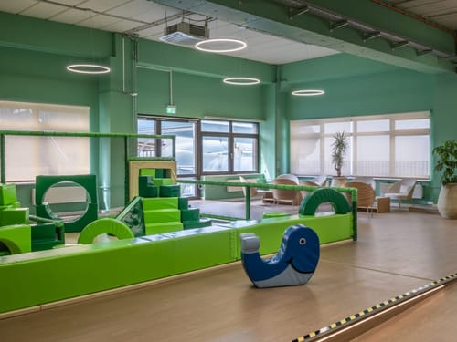Innenansicht eines modernen Indoor-Spielplatzes mit grünen Kletterelementen und einer blauen Wippe in Walform.
