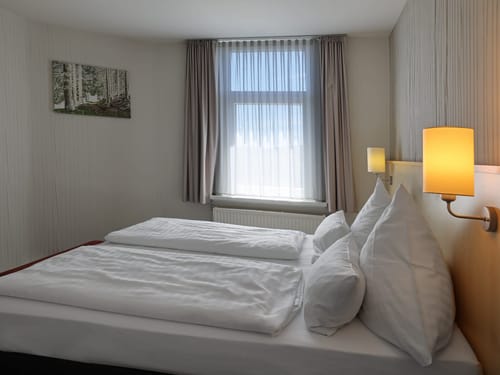 Gepflegtes Doppelzimmer mit weißer Bettwäsche, Wandbeleuchtung und natürlichem Lichteinfall durch ein Fenster mit Vorhängen