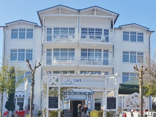 Café Strandkorb vor einem weißen Gebäude mit blauen Akzenten und sonnigem Himmel.