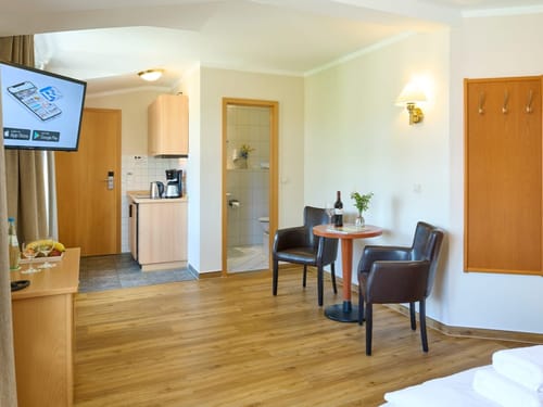 Helles Hotelzimmer mit Holzboden, kleiner Küche, Ecktisch mit zwei Stühlen und modernem Flachbildfernseher.