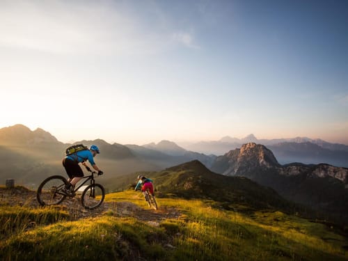 Zwei Mountainbiker fahren bei Sonnenuntergang auf einem Bergpfad.