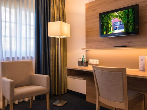Einladendes Hotelzimmer mit modernem Schreibtischbereich, gemütlichem Sessel und Flat-TV an der Holzwand