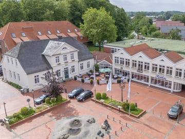 Luftaufnahme eines historischen Gebaeudes und eines modernen Restaurants in einer Stadt mit Kopfsteinpflasterplatz und Springbrunnen.