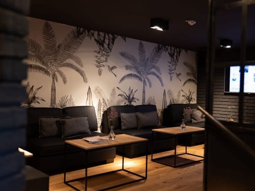 Stilvoll eingerichteter Loungebereich mit schwarzem Sofa und tropischer Wanddekoration.