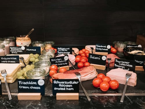Frühstücksbuffet mit einer Auswahl an Aufschnitt, Käse, Hummus, Tomaten und Trauben.