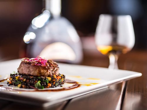 Köstliches Gourmet-Gericht mit edlem Fleisch und Beilage auf einem weißen Teller, begleitet von einem Glaskaraffe edlen Getränks im Hintergrund.