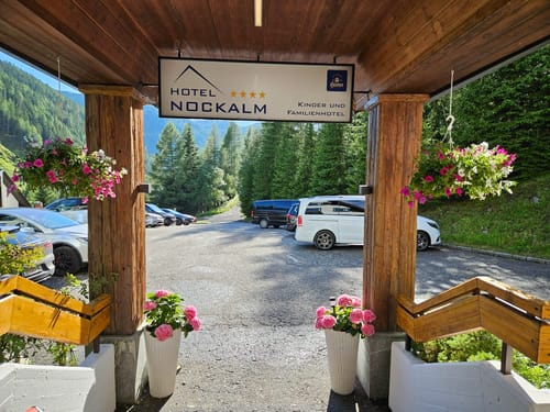Eingang des Hotel Nockalm mit Blick auf den Parkplatz und die umliegenden Berge.