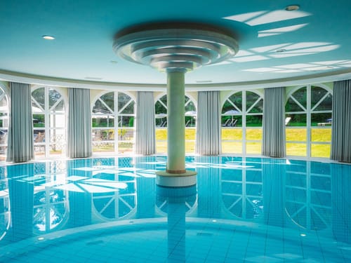 Atemberaubender Innenpool eines Luxushotels mit zentraler Säule, türkisblauem Wasser und eleganten Bogenfenstern mit Gartenblick