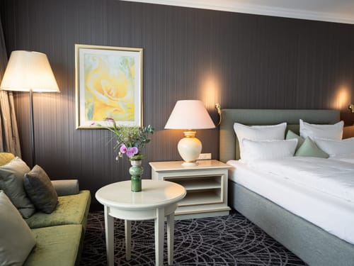 Elegantes Hotelzimmer mit Doppelbett, grünem Samtsessel und dekorativem Blumenstrauß