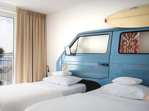 Kreatives Hotelzimmer mit blauem VW-Bus als Bett und Surfbrett an der Wand
