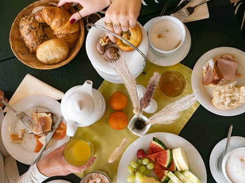 Reichhaltiges Frühstück mit Brötchen, Croissants, frischem Obst und Tee auf einem gedeckten Tisch.