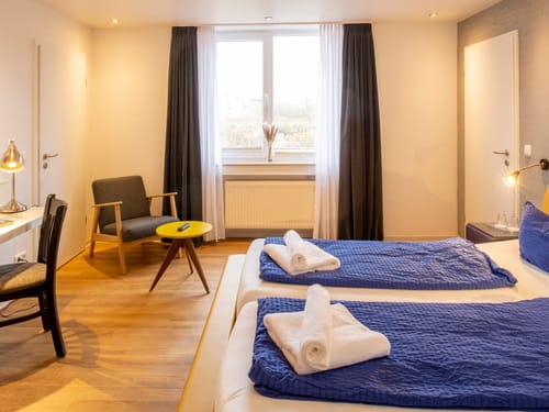 Modern eingerichtetes Hotelzimmer mit zwei Einzelbetten, einem gemütlichen Sessel und Schreibtisch in warmen Farben.