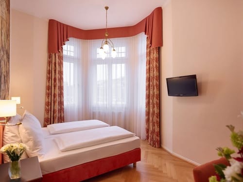 Elegantes Hotelzimmer mit einem Doppelbett, großen Fenstern, einem Fernseher an der Wand und klassischen Vorhängen in warmen Farben.