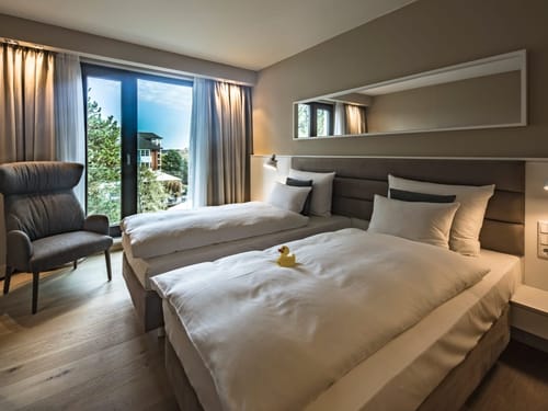 Modernes Hotelzimmer mit zwei Einzelbetten und einem Sessel neben einem großen Fenster.