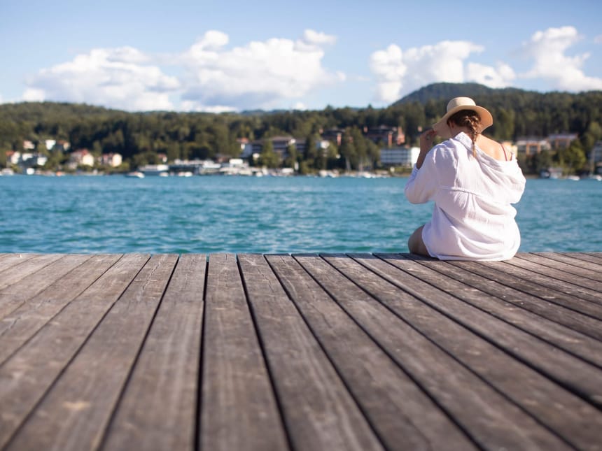 Frau mit Hut sitzt auf einem Steg am See mit Blick auf ein Dorf und bewaldete Hügel im Hintergrund
