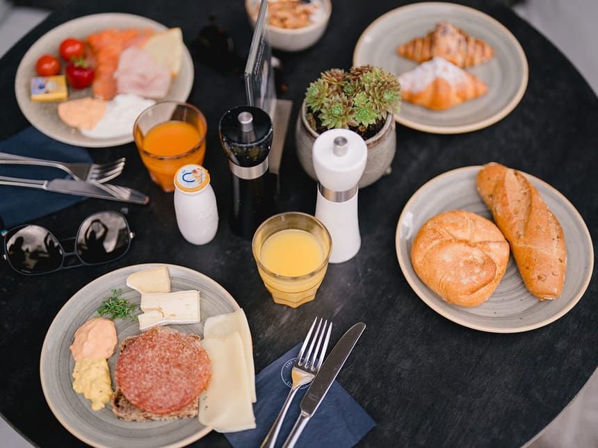 Reichhaltiges Frühstück mit Brötchen, Croissants, Käse, Salami, Räucherlachs, Quark und Orangensaft auf einem Tisch.
