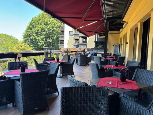 Gemütliche Restaurantterrasse mit Holzsicht, modernen Rattanmöbeln und roten Tischdecken unter einer Markise.