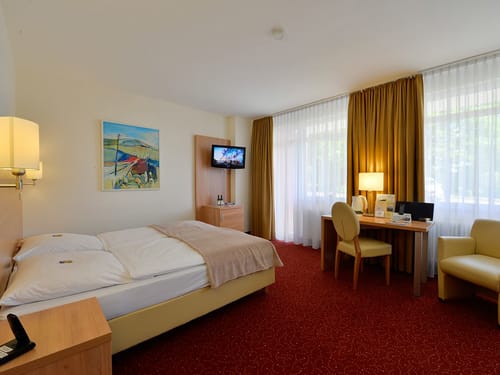 großzügiges Doppelzimmer mit Doppelbett, Schreibtisch, Stuhl, Sessel und TV an der Wand