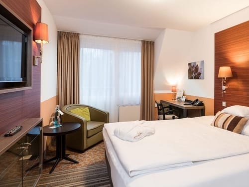 Hotelzimmer in Erdtönen mit TV, Sessel, Tisch und einem Schreibtisch