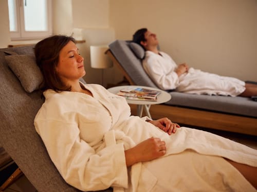 Zwei Personen entspannen im Spa auf Liegen in weißen Bademänteln.