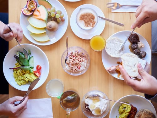 Verschiedene Frühstücksteller mit Obst, Joghurt, Brötchen und Saft auf einem Holztisch.