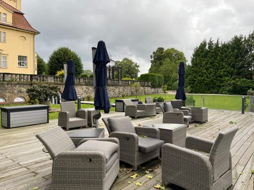 Außenbereich mit gemütlichen Gartenmöbeln und Sonnenschirmen auf einer Terrasse vor einem gelben Gebäude.