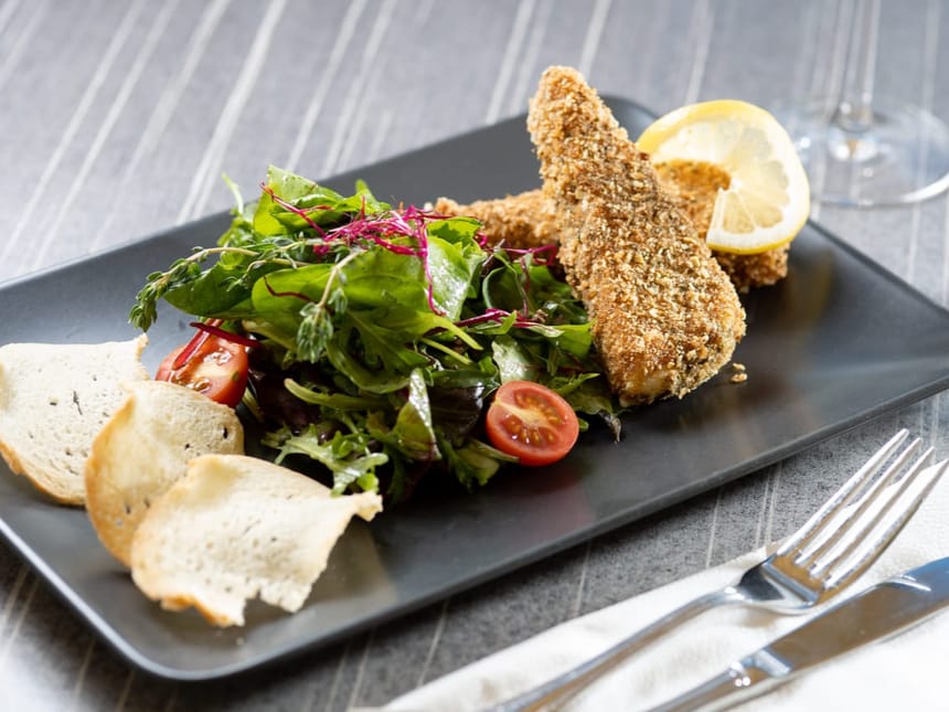 Ein angerichtetes Restaurantgericht auf einem schwarzen rechteckigen Teller: knusprig paniertes Fleisch mit goldbrauner Kruste, begleitet von einem frischen gemischten Blattsalat mit roten Cocktailtomaten, knusprigen Brotscheiben und einer Zitronenscheibe. Das Gericht wird mit Besteck auf einem grauen Tisch serviert und eignet sich perfekt als Vorspeise oder leichtes Hauptgericht.