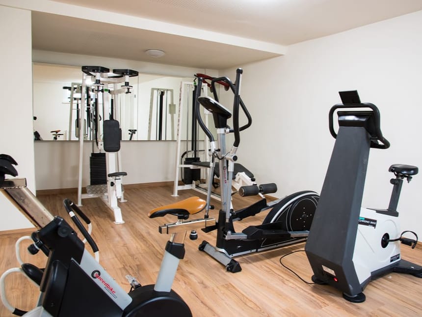 Heller und gut ausgestatteter Fitnessraum mit Holzlaminatboden, großer Spiegelwand und verschiedenen Trainingsgeräten, darunter Fahrradergometer, Crosstrainer und eine Kraftstation für ein effektives Ganzkörpertraining