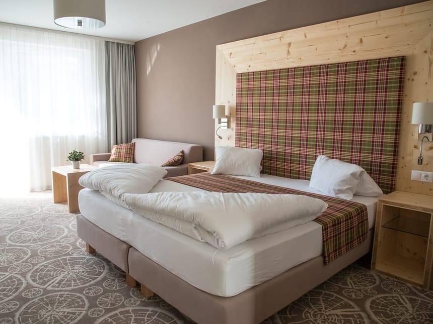 Helles und komfortables Hotelzimmer im alpinen Stil mit einem großzügigen Doppelbett auf Boxspring-Basis, einem markanten Kopfteil aus hellem Zirbenholz mit karierter Polsterung in Rot und Grün, gemustertem Teppich, Holznachttischen mit Wandleuchten sowie einer gemütlichen Sitzecke mit Sofa und natürlichem Lichteinfall durch bodentiefe Fenster