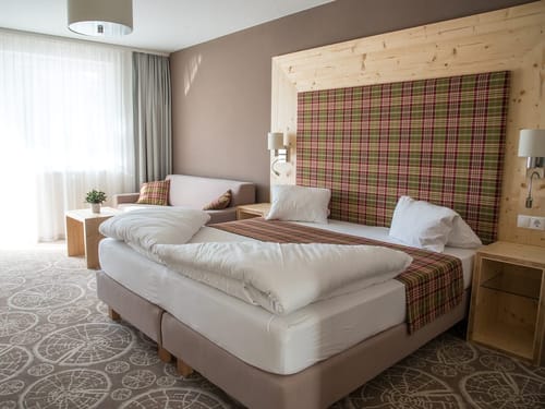 Helles und komfortables Hotelzimmer im alpinen Stil mit einem großzügigen Doppelbett auf Boxspring-Basis, einem markanten Kopfteil aus hellem Zirbenholz mit karierter Polsterung in Rot und Grün, gemustertem Teppich, Holznachttischen mit Wandleuchten sowie einer gemütlichen Sitzecke mit Sofa und natürlichem Lichteinfall durch bodentiefe Fenster