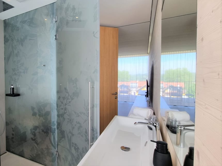 Modernes Hotelbadezimmer mit floraler Glasduschwand, weißem Waschtisch und offenem Blick ins lichtdurchflutete Schlafzimmer
