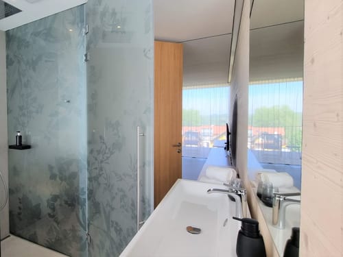 Modernes Hotelbadezimmer mit floraler Glasduschwand, weißem Waschtisch und offenem Blick ins lichtdurchflutete Schlafzimmer