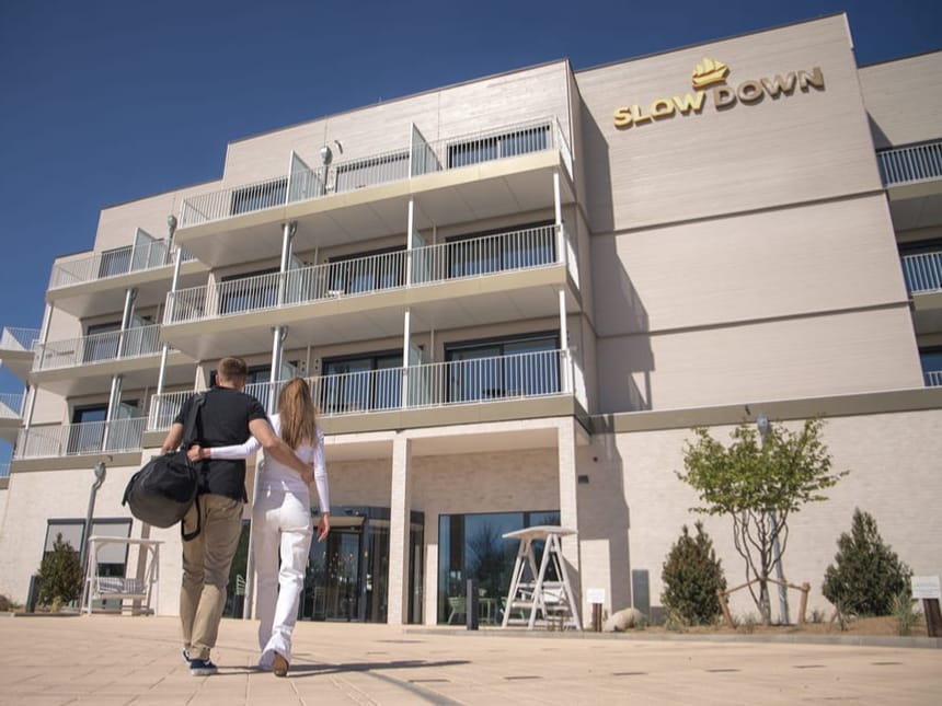 Paar vor dem Eingang eines modernen Hotels mit "Slow Down" Schriftzug.