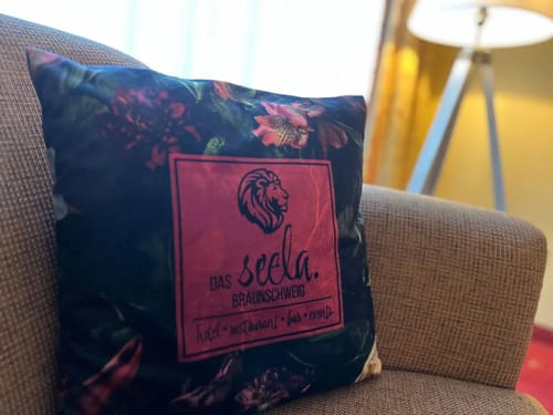 Dekoratives Kissen mit "Das Seela Braunschweig" Motiv auf einem Sessel im Hotelzimmer.