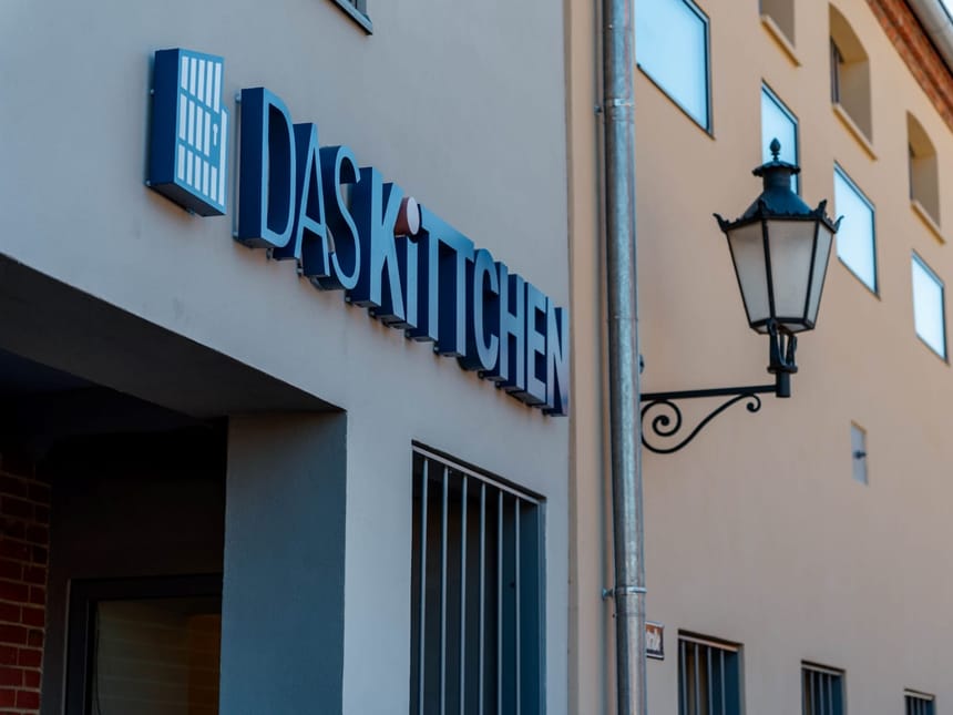 Der Eingangsbereich von DAS KiTCHEN mit dem blauen Markenzeichen an der Fassade und einer dekorativen Straßenlaterne im Vordergrund.