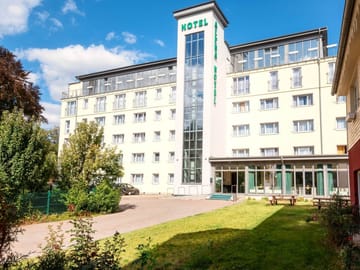 Außenansicht des Alpha Hotels mit grünem Schriftzug, Glasfassade und sonnigem Gartenbereich