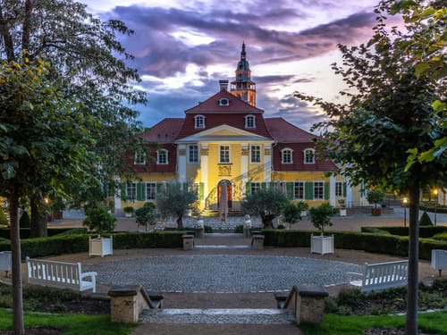 Ein barockes Schloss erstrahlt zur blauen Stunde in warmer Beleuchtung – der gepflegte Schlossgarten mit symmetrischen Wegen und weißen Bänken lädt zum Verweilen ein
