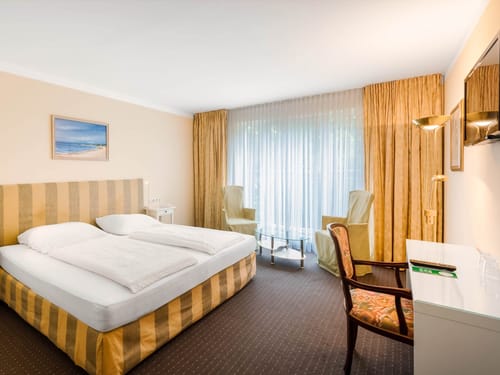 Helles und komfortables Doppelzimmer mit eleganter Einrichtung in warmen Goldtönen, Sitzecke und großem Fensterfront für viel natürliches Licht.