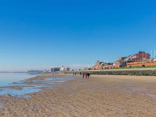 Menschen spazieren an einem sonnigen Sandstrand entlang einer Uferpromenade.