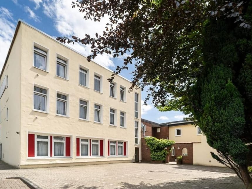 Helles, modernes Gebäude mit mehreren Stockwerken und großen Fenstern, umgeben von Bäumen und einem gepflasterten Hof.