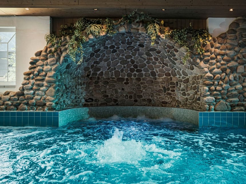 Innenansicht eines Pools mit blauer Kachelumrandung und Steinmauer im Hintergrund, dekoriert mit hängenden Pflanzen.