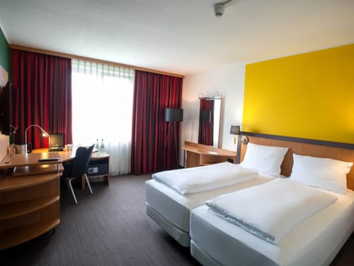Modern eingerichtetes Hotelzimmer mit zwei Einzelbetten, Schreibtisch, und großen Fenstern mit Vorhängen.