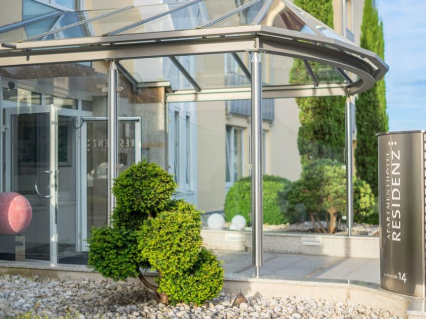 Eingang eines modernen Aparthotels mit Glasüberdachung und gepflegtem Garten.