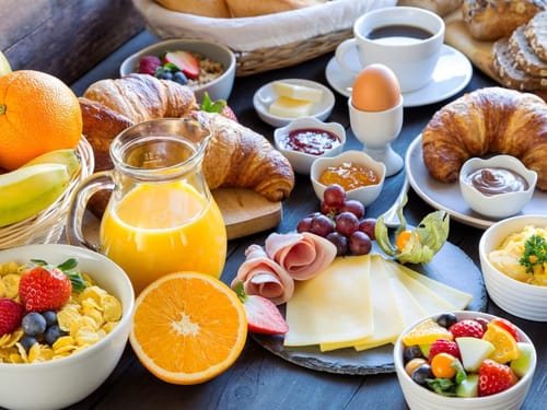 Reichhaltiges Frühstück mit Croissants, Aufschnitt, Käse, frischem Obst, Müsli und Orangensaft.