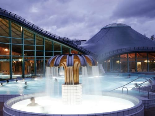 Außenansicht eines Thermalbades bei Nacht mit beleuchtetem Springbrunnen und Badegästen.