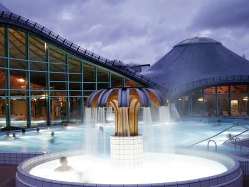 Therme bei Dämmerung mit beleuchteten Pools und Wasserbrunnen